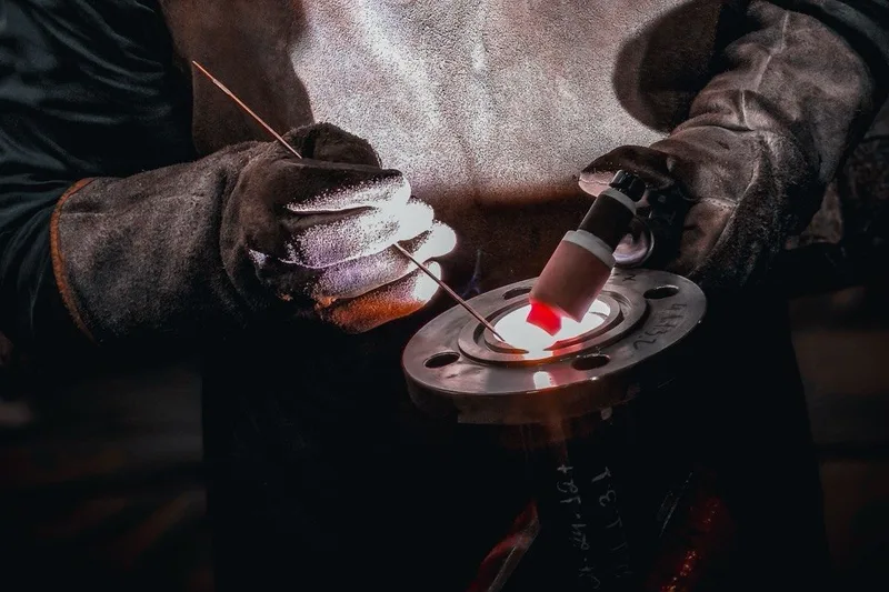 Gloved hands welding a steel flange with a TIG torch.